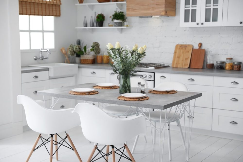 A bright and welcoming kitchen staged for sale, with small upgrades that make a significant impact. The natural light from the window enhances the space, while the modern pendant lights add warmth. The room features a stylish grey concrete dining table with elegant white chairs, and a fresh bouquet centerpiece, illustrating the effect of good lighting and thoughtful touches in staging a home for sale.